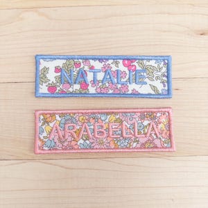 May include: Two embroidered name patches. One patch has a light blue border with the name "NATALIE" in light blue thread. The other patch has a pink border with the name "ARABELLA" in pink thread. Both patches feature a floral pattern.