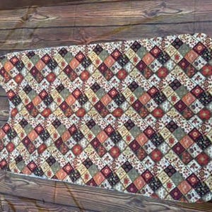 May include: A quilted ironing board cover with a geometric pattern in shades of red, orange, green, and brown. The cover has a curved shape with a pointed end and a padded surface.