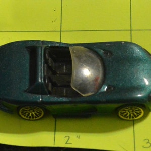 Puede incluir: Un coche de juguete verde oscuro con un parabrisas transparente y un interior negro. El coche tiene ruedas negras con radios amarillos.