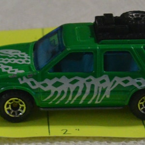 Pode incluir: Carro de brinquedo verde com um design estampado branco e cinza. O carro tem um rack de teto preto com um pneu sobressalente no topo. O carro tem aproximadamente 7,5 cm de comprimento.