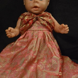 Könnte beinhalten: Eine Porzellanpuppe, die ein rosa-goldenes Kleid mit Schleife trägt. Die Puppe hat ein realistisches Gesicht mit blauen Augen.