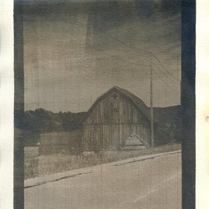 May include: A weathered wooden barn with a gabled roof stands in a field, with a dirt road leading up to it. Power lines run across the sky, and trees line the edge of the field.