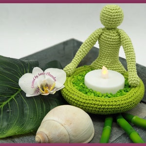 Può includere: Figura verde all'uncinetto che medita con una candela tealight in un portacandele verde all'uncinetto. La figura è circondata da foglie verdi, una conchiglia bianca e bastoncini di bambù.
