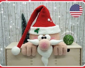 Può includere: Un Babbo Natale all'uncinetto che sporge dal bordo di una scatola di legno. Indossa un cappello rosso con un bordo bianco e una barba bianca. I suoi occhi sono neri e il suo naso è rosa. Lo sfondo è una parete di legno.