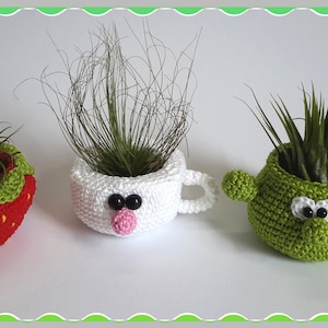 May include: Three crocheted plant pots with cute faces. The pots are shaped like a strawberry, a mug, and a cactus. Each pot has googly eyes and a small mouth. The pots are filled with air plants.