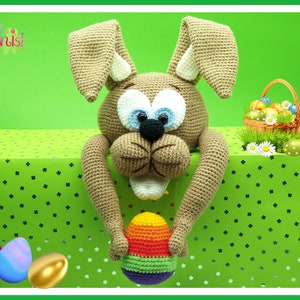 Op de afbeelding: Een gehaakt bruin konijn dat een regenboogkleurig paasei vasthoudt. Het konijn zit op een groene tafelkleed met een wit klaverbladpatroon. Er liggen andere paaseieren op de tafel.