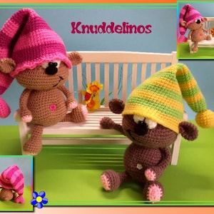 Puede incluir: Dos osos de peluche de crochet marrones que llevan gorros de dormir a rayas rosas y verdes están sentados en un banco blanco. El banco está sobre una superficie de hierba verde con una flor en el fondo. El texto "Knuddelinos" está escrito en el banco.