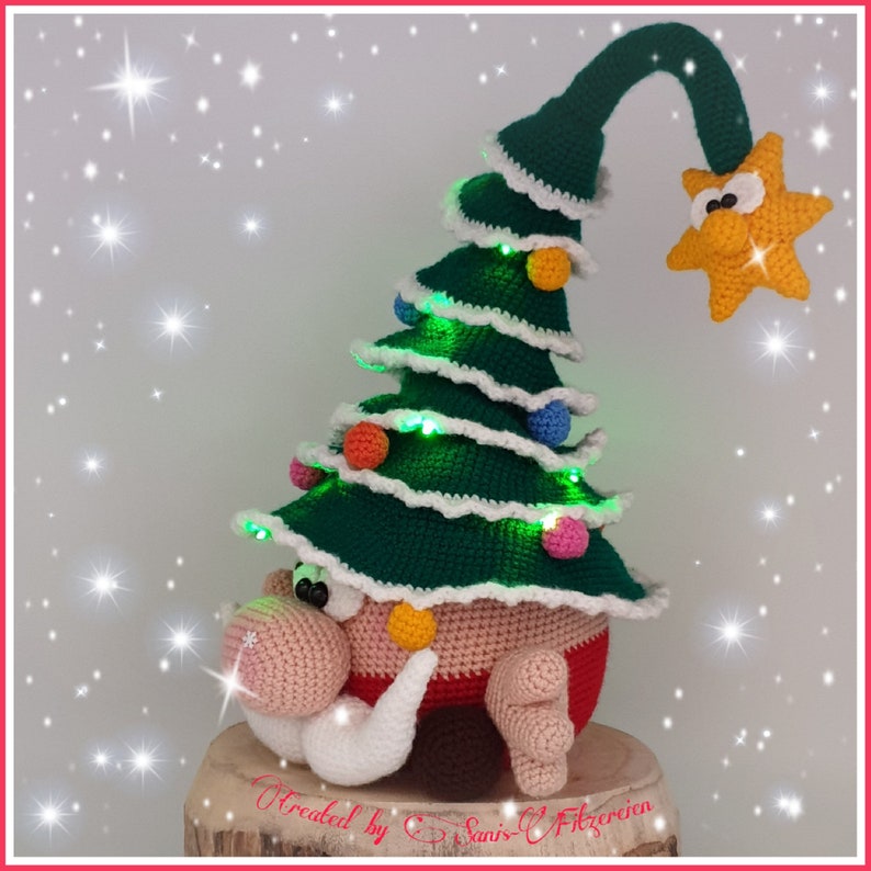 May include: A crocheted gnome wearing a green Christmas tree hat with lights and a yellow star on top. The gnome is sitting on a wooden base.
