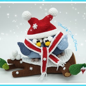 May include: Two blue crocheted birds wearing a red and white scarf and a red Santa hat with a white pom-pom on a brown crocheted branch with green leaves and white snowflakes.