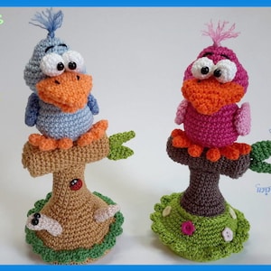 May include: Two crocheted bird figurines perched on tree stumps. The bird on the left is blue with orange beak and feet, the bird on the right is pink with orange beak and feet. Both birds have large white eyes. The tree stumps are brown with green leaves and a worm at the base.