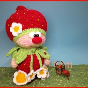 Peut inclure: Une poupée en crochet en forme de fraise portant une cape en feuille verte et un chapeau de fraise rouge avec des fleurs blanches. La poupée tient un panier de fraises et se tient sur une surface herbeuse verte.