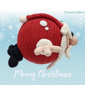 Puede incluir: Un adorno de Papá Noel de crochet en rojo, blanco y negro, con barba blanca y gorro rojo. El adorno está volando por el aire con un fondo azul de copos de nieve. El texto "Feliz Navidad" está escrito debajo del adorno.