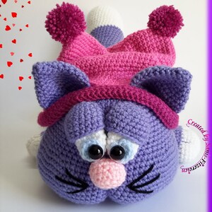May include: A crocheted purple cat wearing a pink hat with white pom-poms. The cat has black eyes, a pink nose, and black whiskers. The text "Created by Sun-Stitcher" is visible in the lower right corner of the image.