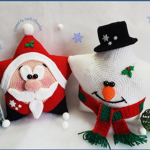 Könnte beinhalten: Zwei gehäkelte sternförmige Kissen, eines zeigt den Weihnachtsmann mit weißem Bart und roter Mütze, das andere zeigt einen Schneemann mit schwarzem Zylinder und grünem und rotem Schal. Die Kissen liegen auf einem weißen Hintergrund mit Schneeflocken.