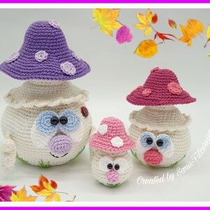May include: Three crocheted mushroom amigurumi toys with different colored hats. The largest mushroom has a purple hat with white polka dots, the middle mushroom has a pink hat with white polka dots, and the smallest mushroom has a pink and white hat with white polka dots. The mushrooms have white bodies with embroidered eyes and mouths.