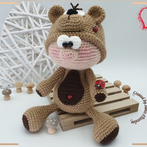 Puede incluir: Un oso de crochet marrón que lleva un sombrero de mariquita y sostiene una mariquita. El oso tiene ojos grandes y redondos y una nariz rosa. El oso está sentado en una tarima de madera.
