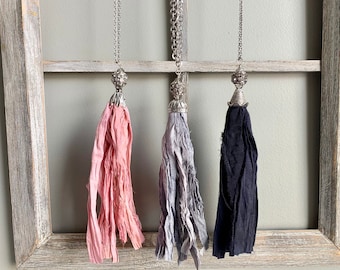 Puede incluir: Tres collares de cadena plateada con borlas de tela. Las borlas son de color rosa, gris y negro.