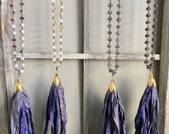 Puede incluir: Cuatro collares con borlas de seda morada que cuelgan de una pantalla de madera. Los collares están hechos de pequeñas cuentas redondas de varios colores, incluyendo blanco, beige y gris.