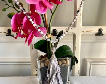 Puede incluir: Un collar de cuentas blancas y grises con un borla gris que cuelga de un dije plateado. El collar está drapeado sobre una planta de orquídea rosa en un jarrón de vidrio.