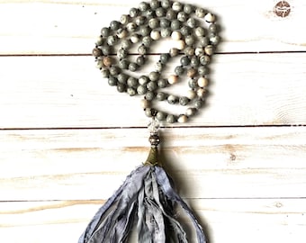 Puede incluir: Un collar mala de piedra gris y blanca con un borla de seda azul. El collar está hecho de cuentas redondas y tiene un acento de metal dorado.