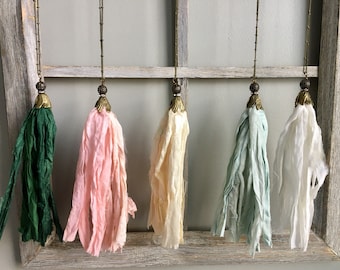 Puede incluir: Cinco collares de borlas de seda con cadenas doradas y tapas metálicas decorativas. Las borlas son en tonos de verde, rosa, crema, azul claro y blanco.
