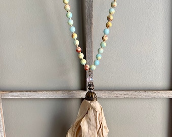 Op de afbeelding: Een lange ketting gemaakt van lichtblauwe, gele en bruine gepatenteerde kralen met een crèmekleurige stoffen kwast die aan een bronzen hanger hangt.