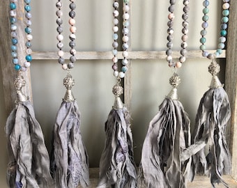 Puede incluir: Cinco collares de borla grises con cuentas plateadas y una borla plateada. Cada collar presenta piedras de diferentes colores, incluyendo azul, rosa y gris.