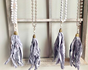 Puede incluir: Cuatro collares con borlas de tela gris y diferentes estilos de cuentas. Las cuentas son blancas, transparentes y marrones.