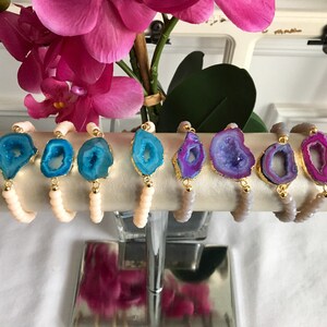 May include: A set of eight agate slice bracelets with gold accents. The bracelets are displayed on a white stand. The agate slices are in shades of blue, purple, and pink.