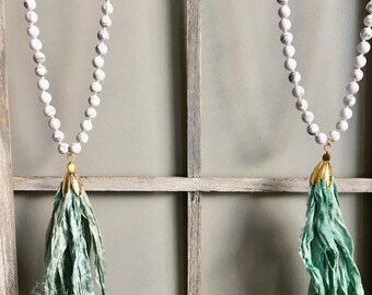Puede incluir: Dos collares de cuentas blancas con colgantes de borla verde que cuelgan de un marco de ventana de madera.