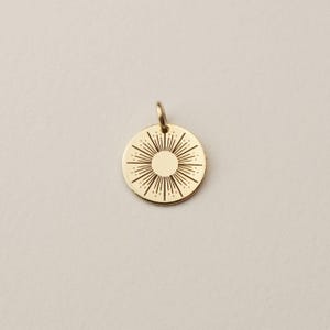 May include: A circular gold pendant featuring an engraved sunburst design with a solid centre circle and radiating lines of varying lengths extending towards the edge. The pendant includes a small attached jump ring at the top for suspension.
