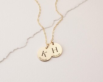 May include: A gold-toned necklace featuring two circular pendants. Each pendant is engraved with a single uppercase letter, K and H, and a numerical date below the letter. The dates are 05.02.07 on the K pendant and 02.15.09 on the H pendant.