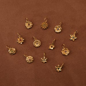 May include: A collection of twelve gold-toned metal flower charms, each featuring a small, uniquely coloured gemstone at its centre. The charms vary in floral shape and petal design, and each includes a small loop at the top for attachment.