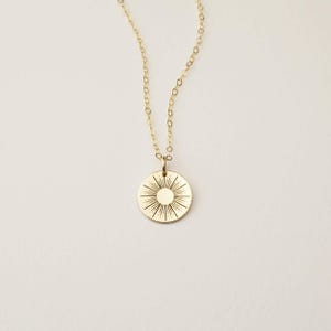May include: A gold-colored necklace featuring a delicate chain and a circular pendant. The pendant displays an engraved sunburst design with a solid center circle surrounded by radiating lines of varying lengths, creating a starburst pattern.