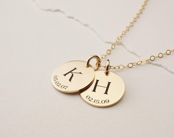 May include: A gold-toned necklace featuring two circular pendants. One pendant is engraved with the letter K and the date 05.02.07, while the other pendant is engraved with the letter H and the date 02.15.09. The pendants hang from a delicate gold chain.