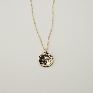 May include: A gold-colored necklace featuring a delicate chain and a small, round pendant. The pendant displays a detailed, dark, high-contrast graphic print of the moon's surface, showing craters and lunar textures against a metallic gold background.