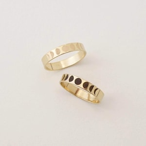 May include: Two gold-coloured metal rings featuring a repeating pattern of moon phases engraved around the band. The top ring displays the moon cycle with subtle indentations, while the bottom ring features dark, high-contrast moon phase details.