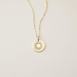 May include: A gold-toned necklace featuring a delicate chain and a circular pendant. The pendant displays an engraved sunburst design with a solid center circle surrounded by radiating lines of varying lengths, creating a star-like pattern.
