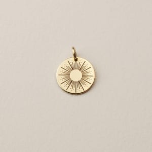 May include: A circular gold pendant featuring a central sunburst design with engraved radiating lines and a small jump ring at the top. The metal surface is polished and reflective, showcasing a detailed starburst pattern etched into the gold disc.