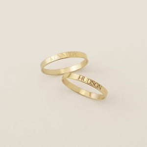 May include: Two thin gold bands with a polished finish. One ring is engraved with the name KENNEDY in a clean serif font, while the other ring features the engraved name HUDSON in the same matching font style, creating a pair of rings.