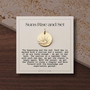 Peut inclure: Un pendentif circulaire doré gravé d'un soleil se levant sur des collines, fixé sur une carte intitulée Suns Rise and Set. La carte contient un texte sur la signification des levers et couchers de soleil et porte la marque KITH & OAK en bas.