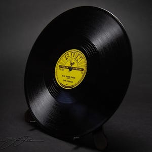 May include: A black vinyl record featuring a yellow Sun Record Company label in the center. The label displays the text Blue Suede Shoes by Carl Perkins. The record is positioned at an angle on a black stand, showcasing its circular grooves.