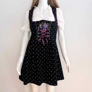 May include: Black dirndl dress with white floral pattern and purple accents. The dress features a square neckline, a bodice with purple lacing, and a white blouse with ruffled sleeves. The dress is displayed on a mannequin.