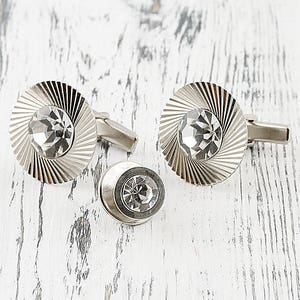 May include: A pair of silver cufflinks with clear crystal accents. The cufflinks have a circular design with a raised center and a textured surface.