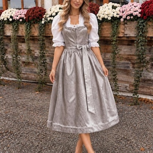 Op de afbeelding: Een grijze dirndl-jurk van gestructureerde stof met een vierkante halslijn met decoratieve rand en een bijpassende ceintuur. De jurk wordt gedragen over een witte blouse met korte pofmouwen en kanten boorden. De rok valt tot op de kuit.