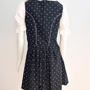 May include: Black dirndl dress with white floral pattern and pink trim. The dress features a fitted bodice, a flared skirt, and a white blouse with ruffled sleeves. The dress is displayed on a mannequin.