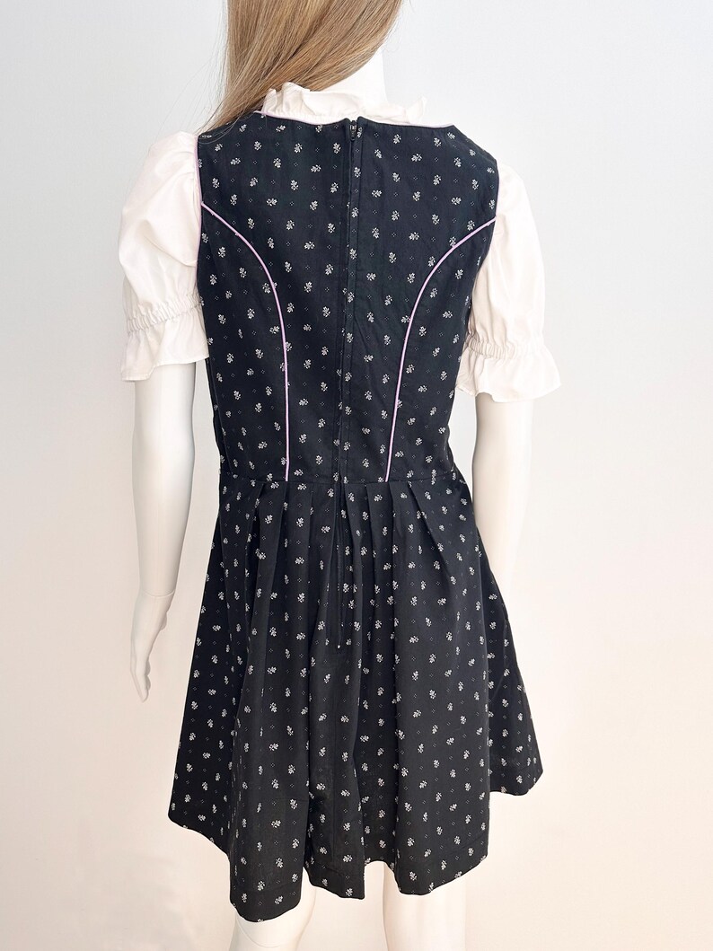 May include: Black dirndl dress with white floral pattern and pink trim. The dress features a fitted bodice, a flared skirt, and a white blouse with ruffled sleeves. The dress is displayed on a mannequin.