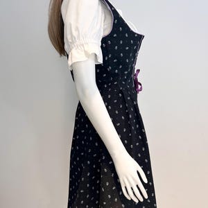 May include: Black dirndl dress with white floral pattern and purple accents. The dress features a fitted bodice with a lace-up detail and a flared skirt. A white blouse with ruffled sleeves is worn underneath.