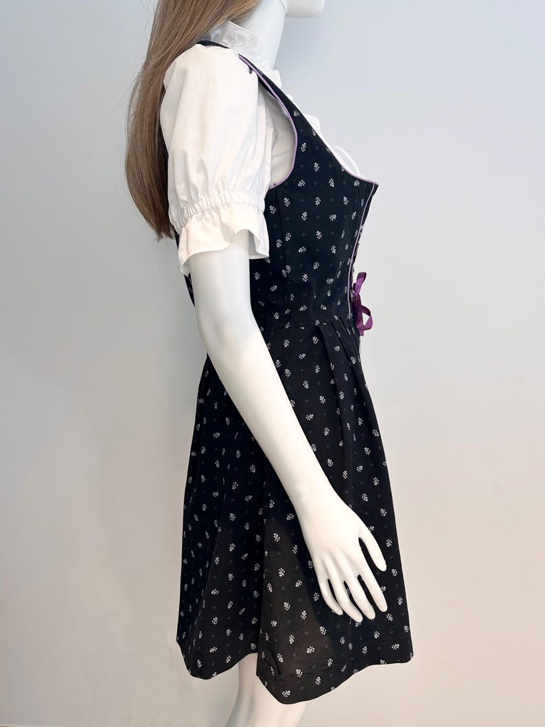 May include: Black dirndl dress with white floral pattern and purple accents. The dress features a fitted bodice with a lace-up detail and a flared skirt. A white blouse with ruffled sleeves is worn underneath.