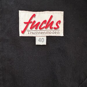 May include: Close-up of a black garment with a white label. The label features the red word "fuchs" in a cursive font, with "Trachtenmoden" below. A smaller tag displays the number "40".
